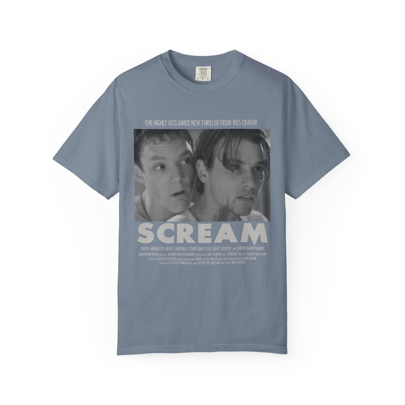 Pu&ograve; includere: Una maglietta blu-grigia con un'immagine in bianco e nero del film "Scream". L'immagine mostra due persone e il titolo "SCREAM" in bianco. Il testo "THE HIGHLY ACCLAIMED NEW THRILLER FROM WES CRAVEN" &egrave; sopra l'immagine.