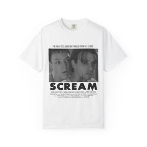 Pu&ograve; includere: T-shirt bianca con un'immagine in bianco e nero del film "Scream". La grafica include il titolo del film e il testo "THE HIGHLY ACCLAIMED NEW THRILLER FROM WES CRAVEN". La maglietta elenca anche i nomi degli attori.