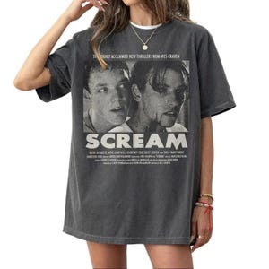 Pu&ograve; includere: T-shirt grigio scuro con un design di locandina del film "Scream". Il design include il titolo del film e le immagini di due attori. La maglietta include anche il testo "HIGHLY ACCLAIMED NEW THRILLER FROM WES CRAVEN."