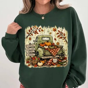 Puede incluir: Una sudadera de cuello redondo verde oscuro con un estampado gráfico de un camión vintage cargado de calabazas. El diseño incluye un poste de madera con el texto PUMPKINS, APPLE CIDER, HAYRIDES, BONFIRES, además de hojas de otoño y una taza.