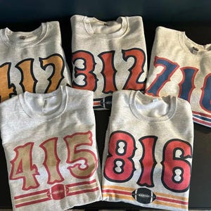 May include: Five light grey sweatshirts, each displaying a different area code in large, bold numbers. The numbers are in various colours, including orange, red, and blue. Each sweatshirt features a football graphic at the bottom.