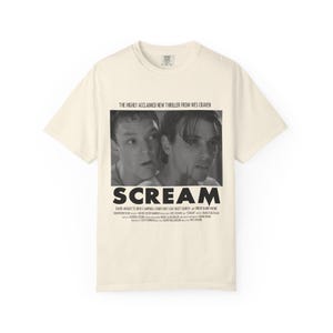 Pu&ograve; includere: T-shirt color crema con immagine in bianco e nero del film "Scream". La grafica include il titolo del film e il testo "THE HIGHLY ACCLAIMED NEW THRILLER FROM WES CRAVEN". Sotto il titolo sono elencati i nomi degli attori.