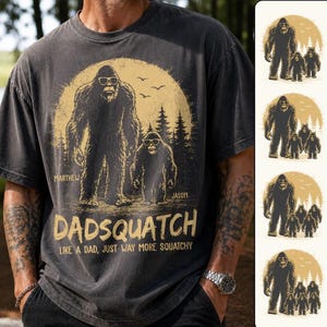 May include: A charcoal gray t-shirt featuring a graphic of Bigfoot figures wearing sunglasses in front of a large yellow moon and forest. The text reads DADSQUATCH LIKE A DAD, JUST WAY MORE SQUATCHY, with names Marthew and Jason printed nearby.