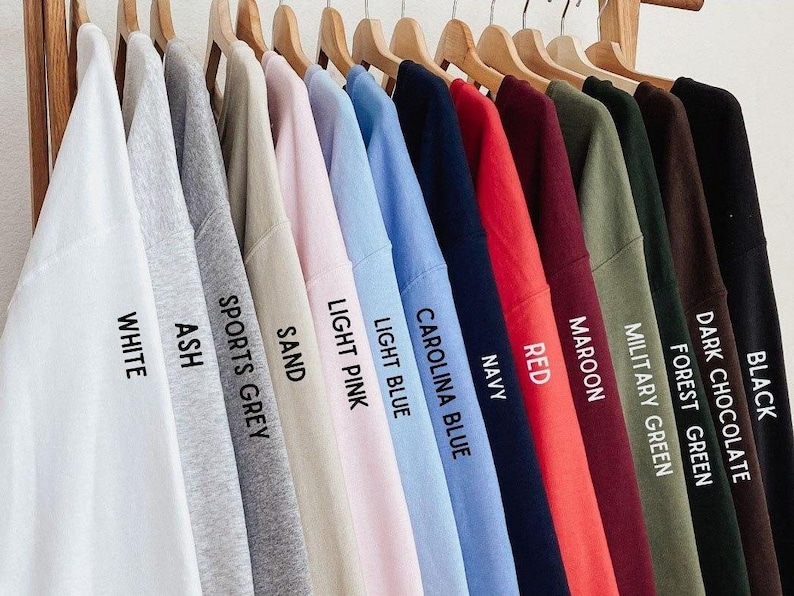 Pu&ograve; includere: Un'esposizione di felpe di vari colori appese a grucce di legno. I colori includono bianco, cenere, grigio sportivo, sabbia, rosa chiaro, azzurro, Carolina blu, blu navy, rosso, bordeaux, verde militare, verde foresta, cioccolato fondente e nero. Ogni felpa ha un'etichetta di colore.