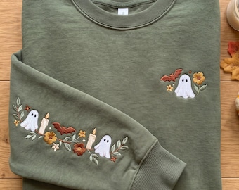 Pode incluir: Uma camisola de gola redonda verde-azeitona com detalhes bordados. O peito apresenta um pequeno fantasma, um morcego e uma flor. A manga apresenta um padrão de videira bordado maior com dois fantasmas, duas velas, um morcego e várias flores.