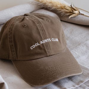 以下が含まれることがあります： 「COOL AUNTS CLUB」の文字が白で刺繍された茶色の野球帽。帽子は柔らかい生地で作られており、つばがカーブしています。帽子はニュートラルカラーの布の上に置かれています。