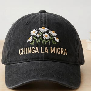 May include: Black baseball cap with embroidered white daisies and the text "CHINGA LA MIGRA" in gold thread. The cap has a curved brim and a button on top. A casual accessory.