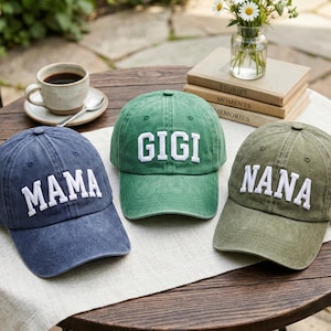 May include: Three baseball caps in navy blue, green, and olive green, each with white embroidered text. The caps read "MAMA", "GIGI", and "NANA". The caps are displayed on a wooden table with a cup of coffee and a small vase of flowers.