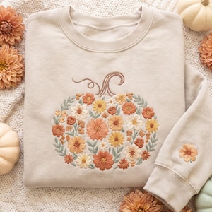 May include: A beige crewneck sweatshirt featuring a large central embroidery of a pumpkin composed of various orange, yellow, and white flowers with green leaves. A smaller matching floral embroidery is visible on the sleeve cuff.