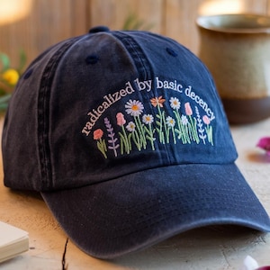 以下が含まれることがあります： 刺繍の花柄と「radicalized by basic decency」の文字が入ったネイビーブルーのベースボールキャップ。キャップはカーブしたつばとクラシックな6パネルデザインが特徴です。刺繍には、ピンク、オレンジ、白の色合いの花が様々含まれています。