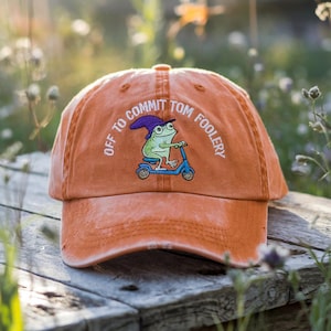 Op de afbeelding: Oranje baseballpet met de geborduurde tekst "Off to Commit Tom Foolery" en een kikker op een scooter. De pet heeft een verweerde uitstraling en een gebogen klep.