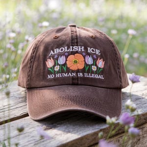 May include: Brown baseball cap with embroidered floral design and the text "ABOLISH ICE" and "NO HUMAN IS ILLEGAL". The cap features a curved brim and adjustable strap, suitable for casual wear.
