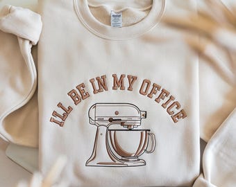 May include: Cream-colored sweatshirt with the embroidered text "I'LL BE IN MY OFFICE" above a brown and cream-colored stand mixer. The sweatshirt is folded, with the sleeves visible on either side.