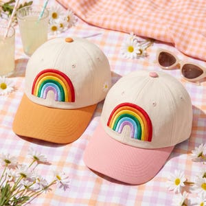 May include: Two baseball caps with off-white crowns and embroidered rainbow designs on the front. One cap features an orange brim and a small white daisy on the side, while the other cap has a pink brim and a matching small white daisy on the side.