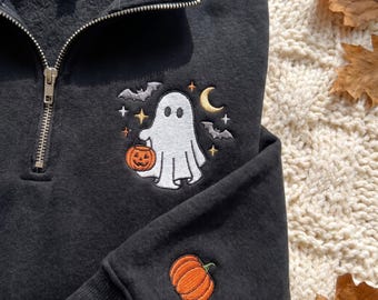 Pode incluir: Uma camisola preta com fecho curto, apresentando um design bordado de um fantasma branco a segurar uma abóbora laranja, rodeado por morcegos cinzentos, uma lua crescente amarela e pequenas estrelas. Uma abóbora adicional está bordada na manga.