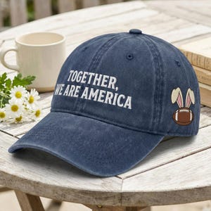 May include: A navy blue baseball cap with the text "TOGETHER, WE ARE AMERICA" embroidered in white. The cap features a football with bunny ears on the side. The hat is sitting on a wooden table.