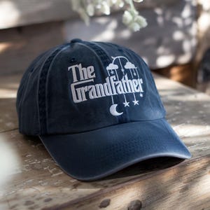 May include: Navy blue baseball cap with the embroidered text "The Grandfather" in white. The design includes clouds, stars, and a crescent moon. A stylish accessory for any grandfather.