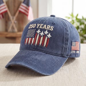 May include: A blue baseball cap with the text "250 YEARS" above an embroidered American flag design with jets. The cap also features the text "1776-2026" and a small American flag on the side. The cap is made of a soft, worn material.