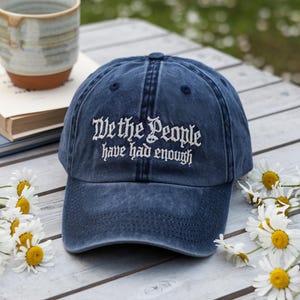 May include: A navy blue baseball cap with the embroidered text "We the People have had enough." The hat is sitting on a white wooden surface, surrounded by daisies and a ceramic mug.