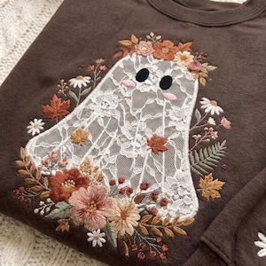 May include: A dark brown sweatshirt featuring an embroidered ghost figure made of white lace. The ghost is surrounded by an intricate floral arrangement including orange, pink, and white flowers, green leaves, and small autumn-colored maple leaves.