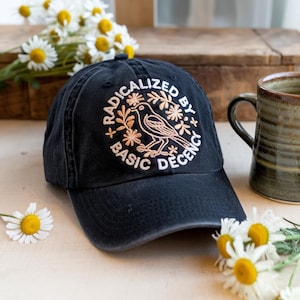 May include: A navy blue baseball cap with the embroidered text "RADICALIZED BY BASIC DECENCY" in white and peach. The design includes a bird and floral elements. Daisies and a mug are in the background.