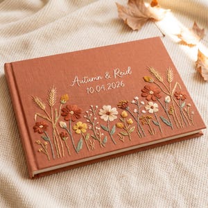 May include: A rectangular photo album with a burnt orange fabric cover featuring embroidered wildflowers in shades of rust, yellow, cream, and sage green. The text "Autumn & Reid" and the date "10.04.2026" are embroidered in white on the front cover.
