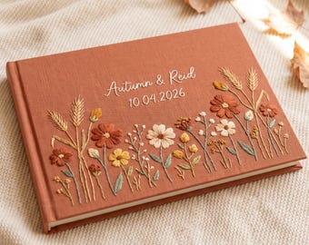 Puede incluir: Un álbum de fotos rectangular con una cubierta de tela color naranja quemado que presenta flores silvestres bordadas en tonos óxido, amarillo, crema y verde salvia. El texto "Autumn & Reid" y la fecha "10.04.2026" están bordados en blanco.
