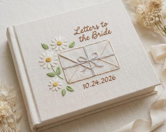 Pode incluir: Um livro quadrado revestido de tecido branco-sujo com detalhes bordados na capa. O bordado inclui o texto "Letters to the Bride", a data "10.24.2026", uma ilustração de um envelope com um laço e flores de margarida brancas.