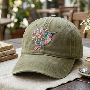 May include: Olive green baseball cap featuring an embroidered hummingbird design. The hummingbird is depicted with colorful wings and a long beak, surrounded by floral accents. The cap has a curved brim and a button on top.