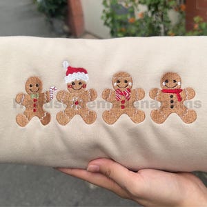 Puede incluir: Una sudadera beige con cuatro hombres de jengibre bordados. Cada hombre de jengibre tiene una cara sonriente y está decorado con una bufanda, un bastón de caramelo o un gorro de Papá Noel. El diseño es festivo y temático de las fiestas.