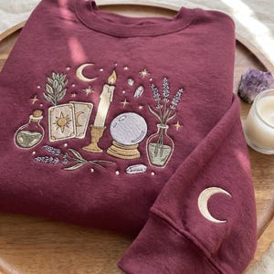 May include: A maroon crewneck sweatshirt featuring intricate embroidery of a candle, a crystal ball, tarot cards, a potion bottle, and lavender sprigs. The design includes celestial stars and moons, with a crescent moon embroidered on the sleeve cuff.