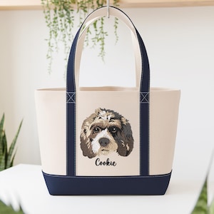 May include: Cream-colored tote bag with a navy blue base and handles. The bag features an embroidered portrait of a dog with brown, black, and white fur. The dog's name, "Cookie," is embroidered below the portrait.