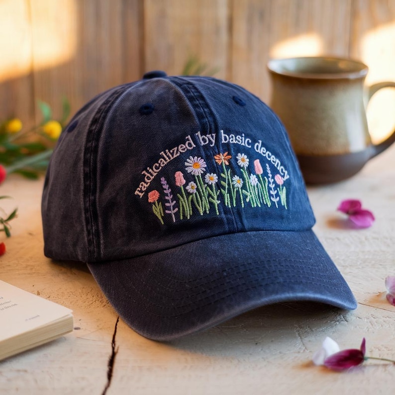Washed navy blue baseball cap with "radicalized by basic decency" embroidered in white above wildflower illustrations, laid on a wooden surface with soft petals and a ceramic mug in the background