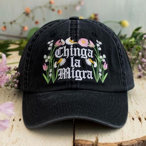 May include: Black baseball cap with embroidered floral design and the text "Chinga la Migra" in white gothic lettering. The floral embroidery includes pink tulips, white daisies, and green leaves. The cap is made of a washed, faded black fabric.