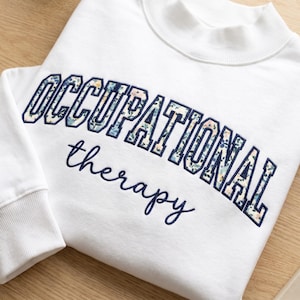 May include: A white long-sleeve sweatshirt with a mock neck collar. The word OCCUPATIONAL is embroidered in a collegiate font with a floral pattern fill, and the word therapy is embroidered below in a dark blue cursive script font.