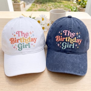 May include: Two baseball caps, one white and one navy blue, are displayed. Both caps feature the embroidered text "The Birthday Girl" in colorful, playful lettering. The white cap has a rainbow color scheme, while the navy cap has a pink, orange, and green color scheme.
