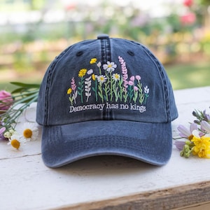 May include: A navy blue baseball cap with embroidered wildflowers in yellow, white, and pink. The text "Democracy has no kings" is embroidered below the flowers. The cap is on a white wooden surface.