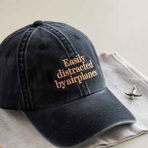 May include: A black baseball cap with the embroidered text "Easily distracted by airplanes" in a tan color. The cap is made of a washed, faded fabric and has a curved brim. A small airplane pin rests on a folded cloth.