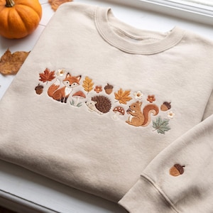 May include: A beige crewneck sweatshirt featuring a central horizontal embroidered design of a fox, a hedgehog, and a squirrel surrounded by autumn leaves, mushrooms, and acorns. A single acorn is embroidered on the lower right sleeve cuff.