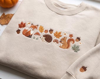 Pode incluir: Uma camisola de gola redonda bege com um bordado horizontal central de uma raposa, um ouriço e um esquilo rodeados por folhas de outono, cogumelos e bolotas. Uma única bolota está bordada no punho da manga direita.