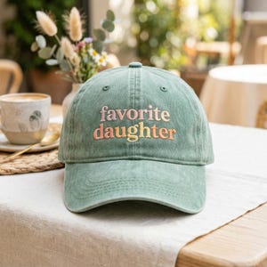 K&ouml;nnte beinhalten: Eine salbeigr&uuml;ne Baseballkappe mit dem gestickten Text "favorite daughter" in Rosa und Gelb. Der Hut ist aus einem weichen, abgenutzten Material gefertigt und hat einen gebogenen Schirm. Die Kappe liegt auf einer wei&szlig;en Tischdecke.