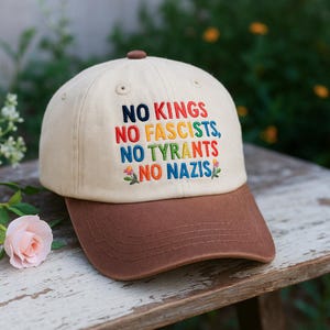May include: A beige baseball cap with a brown brim. The cap features embroidered text in multiple colors that reads "NO KINGS, NO FASCISTS, NO TYRANTS, NO NAZIS". The cap is sitting on a wooden surface.