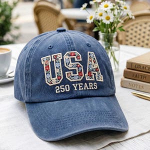 Puede incluir: Gorra de b&eacute;isbol azul con la palabra "USA" bordada con un dise&ntilde;o floral. El texto "250 YEARS" est&aacute; bordado debajo. La gorra est&aacute; sobre una superficie blanca.