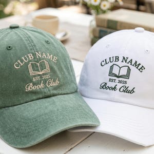 May include: Two baseball caps, one sage green and one white, are displayed. Both caps feature the embroidered text "CLUB NAME" above an open book graphic, with "EST. 2025" and "Book Club" below. The caps are on a white surface.