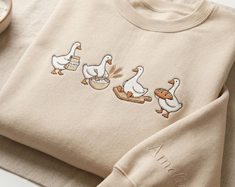 May include: Beige sweatshirt featuring an embroidered design of four geese in a baking scene. The geese are depicted carrying flour, mixing ingredients, and holding a loaf of bread. The sleeve cuff has the name "Amelia" embroidered in a script font.