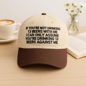 May include: A beige and brown baseball cap with the text "IF YOU'RE NOT DRINKING 13 BEERS WITH ME I CAN ONLY ASSUME YOU'RE DRINKING 13 BEERS AGAINST ME" embroidered in black. The cap has a curved brim and a button on top.