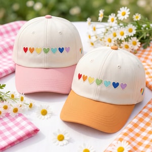 May include: Two baseball caps with a cream-colored crown and a colored brim. Each cap features a row of embroidered rainbow hearts. One cap has a pink brim, the other an orange brim. A small rainbow is embroidered on the side of the orange brim cap.