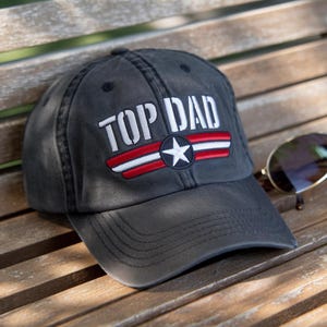 May include: A charcoal gray baseball cap with the text "TOP DAD" in white block letters. The cap features a red and white striped emblem with a white star. Sunglasses rest on a wooden surface next to the cap.