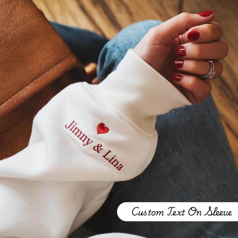 May include: White sweatshirt sleeve with the embroidered text "Jimmy & Lina" and a red heart. The text "Custom Text On Sleeve" is at the bottom. The sleeve is ribbed at the cuff.