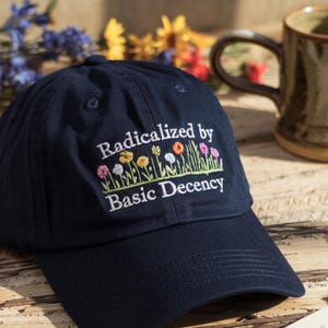 Op de afbeelding: Een marineblauwe baseballpet met witte borduurwerk met de tekst "Geradicaliseerd door elementaire fatsoen". De borduurwerk bevat ook een rij kleurrijke bloemen.